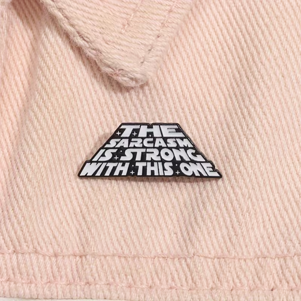 A close-up of a light pink textured fabric, likely a denim jacket or sweatshirt, featuring a black and white triangular enamel pin with the text 'THE SARCASM IS STRONG WITH THIS ONE'.