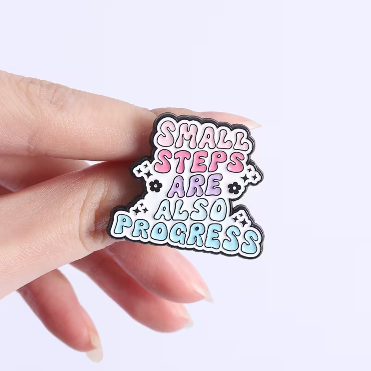 A hand holding a small enamel pin that reads 'SMALL STEPS ARE ALSO PROGRESS' in pastel pink, purple, and blue lettering with black outlines on a light background.