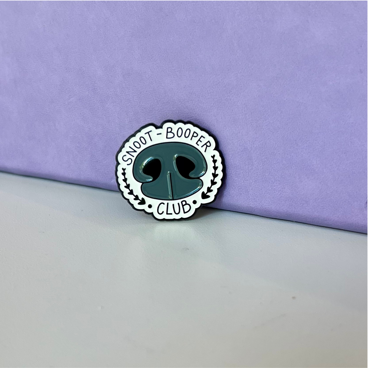 A small enamel pin shaped like a dog nose with the words 'SNOOT - BOOPER CLUB' around it, placed on a white surface against a lavender background.