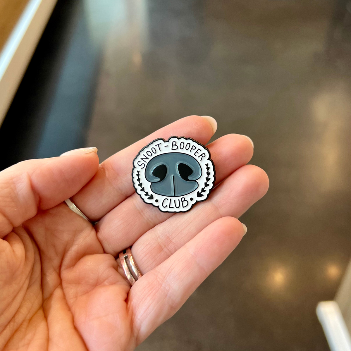 A hand holding a small enamel pin shaped like a dog nose with the words 'SNOOT-BOOPER CLUB' around it, photographed against an indoor blurred background.