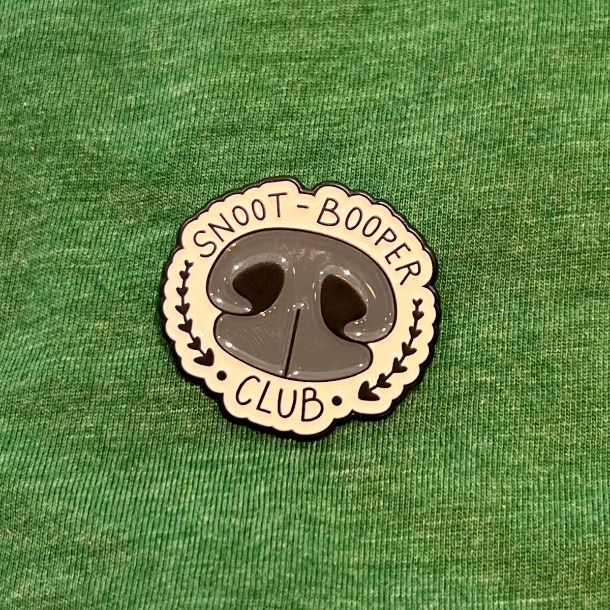 A circular enamel pin shaped like a dog nose with the words 'SNOOT-BOOPER CLUB' around it, placed on a textured green fabric background.