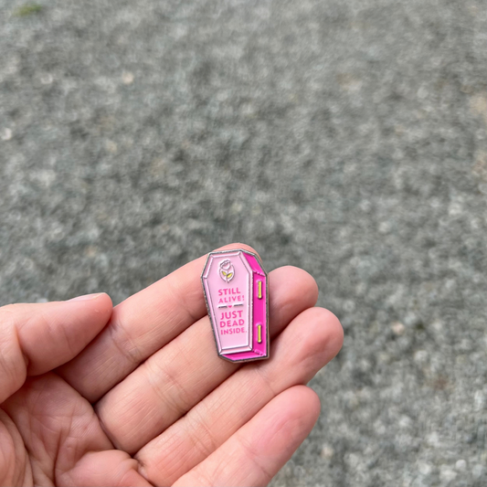 A hand displays the Still Alive! Just Dead Inside Pin by RainBee Designs LLC—a small pink coffin-shaped enamel accessory featuring the phrase “STILL ALIVE JUST DEAD INSIDE”—against a softly blurred gravel background.