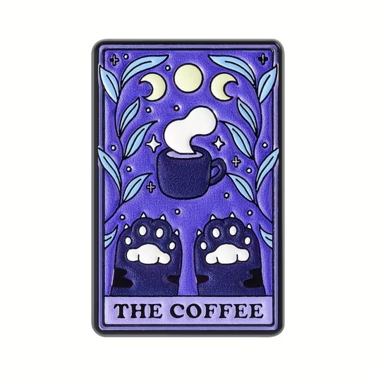 A rectangular enamel pin or sticker featuring a stylized tarot card design titled 'THE COFFEE'. The artwork shows a dark coffee cup with steam, crescent moons, stars, and two paw prints at the bottom, all on a purple background with light blue leaves.