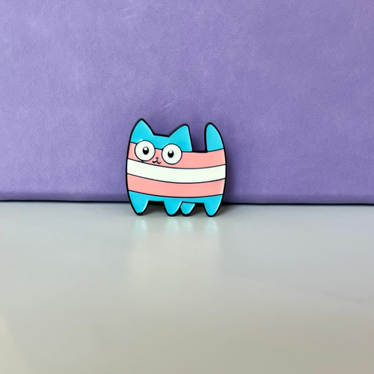 A small enamel pin shaped like a cartoon cat with turquoise blue legs and ears, pink and white horizontal stripes, and large white eyes with black outlines, standing on a light surface against a purple backdrop.