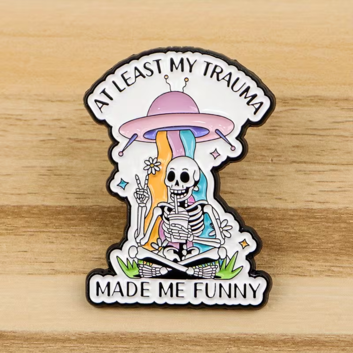 A die-cut enamel pin featuring a cartoon skeleton sitting under a pink UFO with rainbow stripes, holding flowers and making a peace sign. The top text reads 'AT LEAST MY TRAUMA' and the bottom reads 'MADE ME FUNNY'.