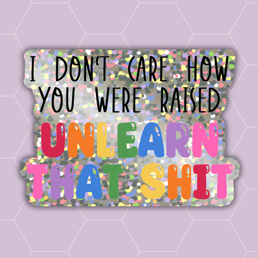A die-cut sticker with a glittery multicolor background and bold black text reading 'I DON'T CARE HOW YOU WERE RAISED' followed by large colorful letters spelling 'UNLEARN THAT SHIT' in red, orange, yellow, green, blue, purple, and pink. The sticker is centered on a light lavender background with a faint hexagonal pattern.