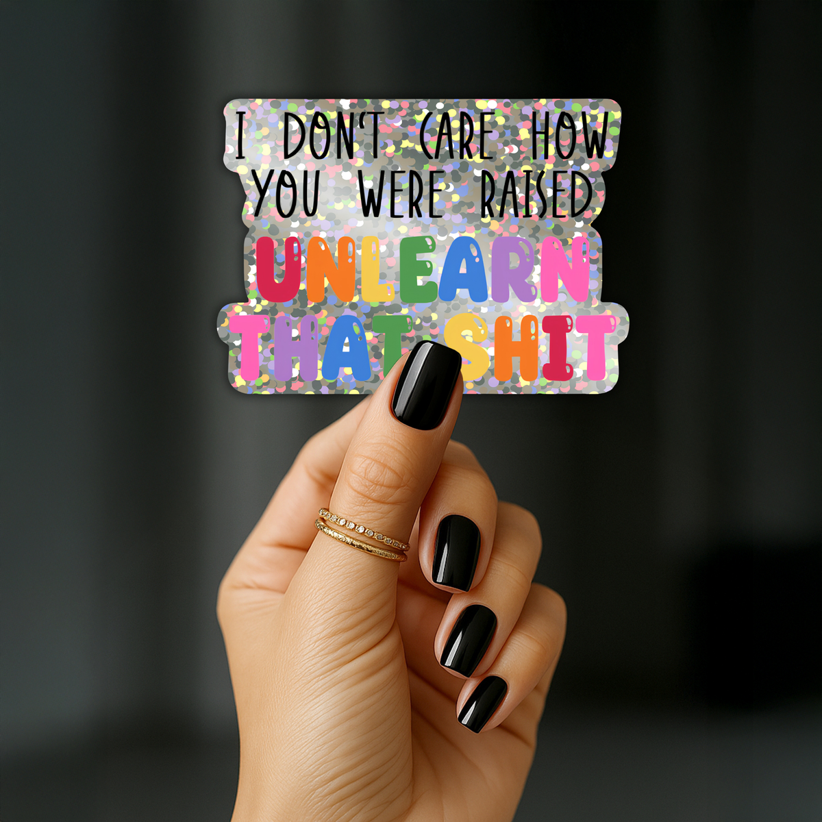 A hand with black nail polish holds a rectangular sticker with a holographic glitter background and the text 'I DON'T CARE HOW YOU WERE RAISED UNLEARN THAT SHIT' in colorful letters.