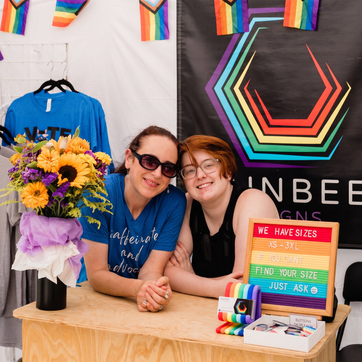 Two women posing behind a wooden table at what appears to be a booth or event. A rainbow-striped banner hangs behind them, and a sign on the table reads 'WE HAVE SIZES XS - 3XL.