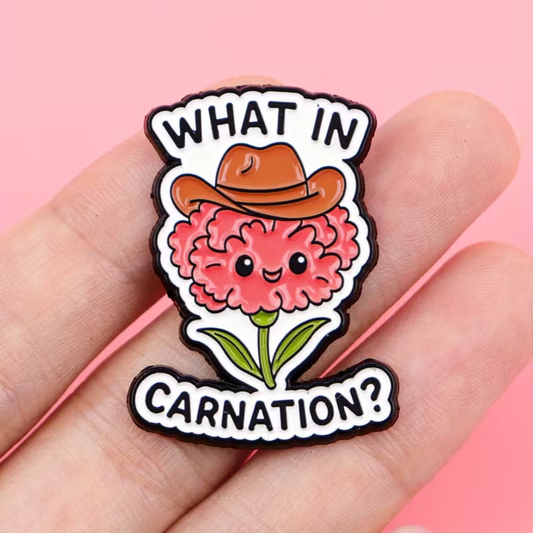 A close-up of a hand holding a cartoon enamel pin shaped like a pink carnation flower wearing a brown cowboy hat, with the text 'WHAT IN CARNATION?' on a pink background.