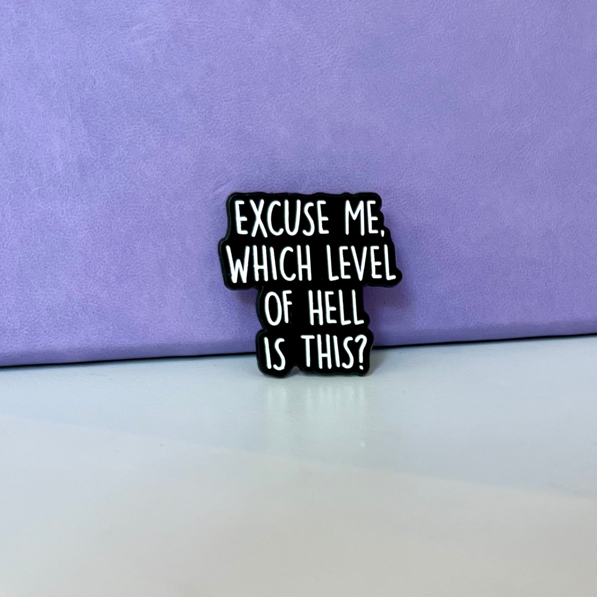 A small black enamel pin with white text reading 'EXCUSE ME, WHICH LEVEL OF HELL IS THIS?' standing on a white surface against a purple background.