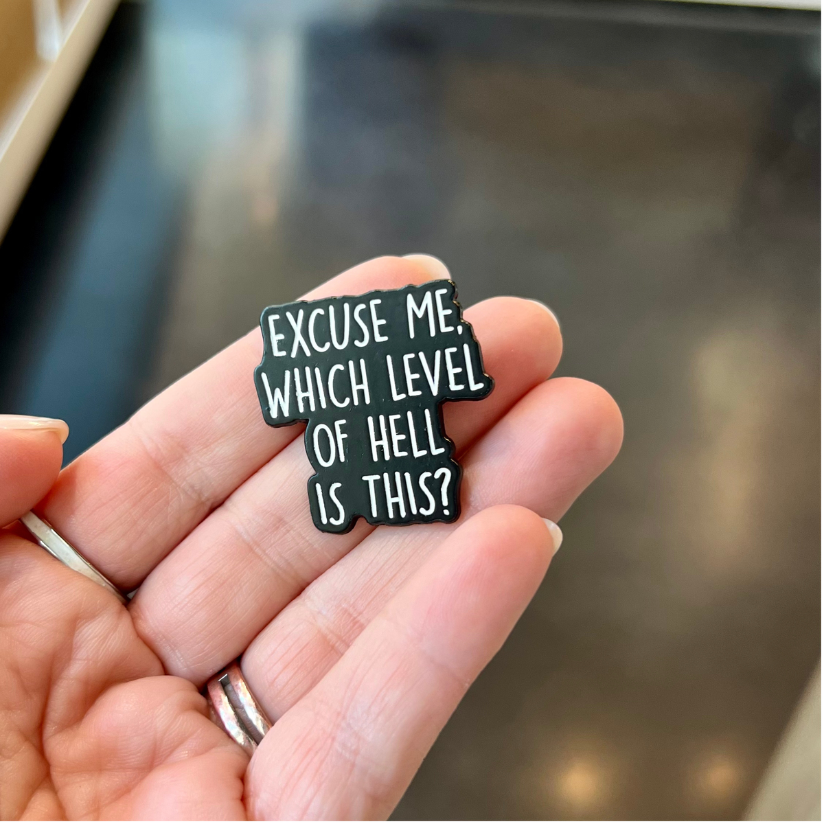 A hand holding a small black enamel pin with white text that reads 'EXCUSE ME, WHICH LEVEL OF HELL IS THIS?' against a blurred dark background.