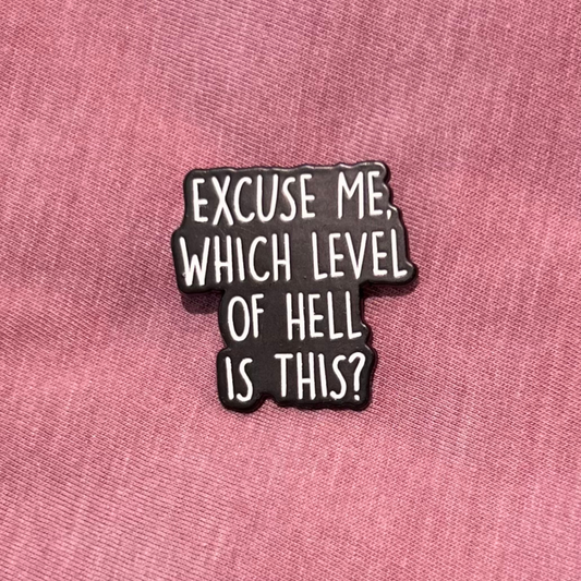 A black enamel pin with white text reading 'EXCUSE ME, WHICH LEVEL OF HELL IS THIS?' displayed on a textured pink fabric background.