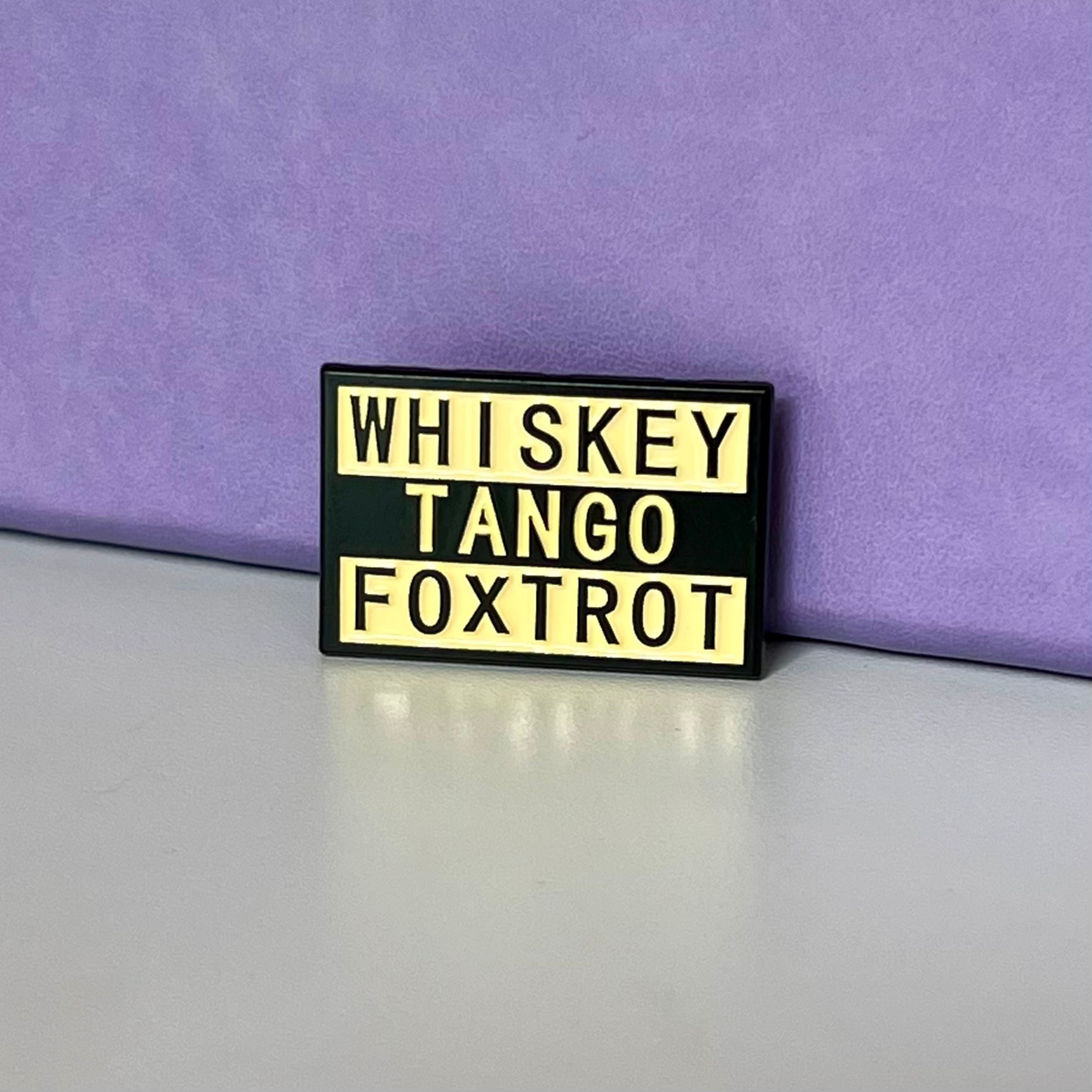 A rectangular enamel pin with a black background and pale yellow lettering that reads 'WHISKEY TANGO FOXTROT', photographed on a light gray surface against a purple backdrop.