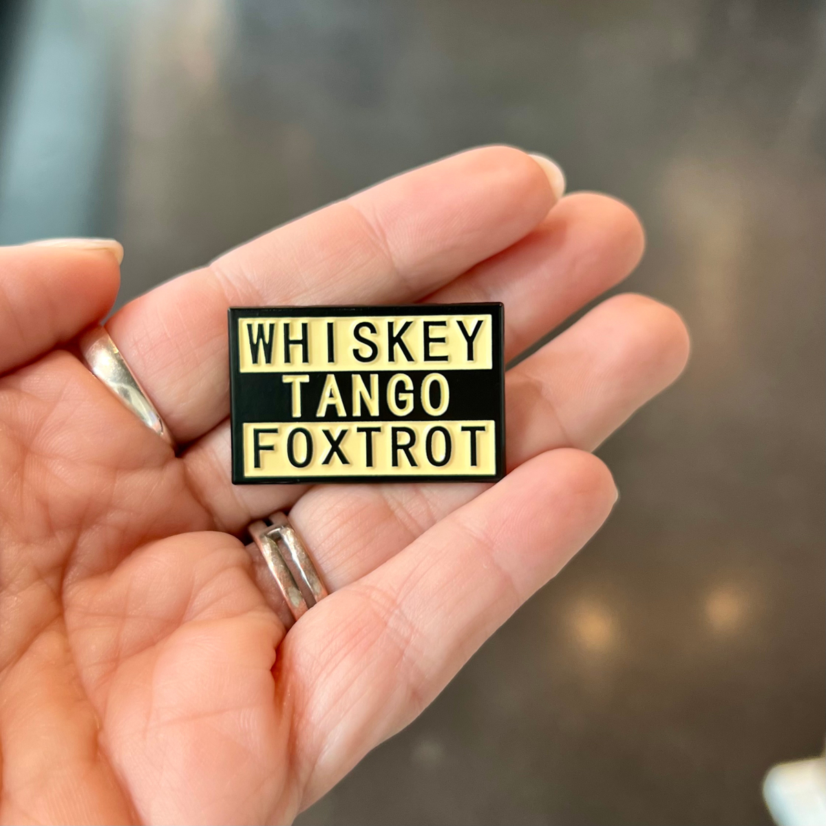 A hand holding a small rectangular enamel pin that reads 'WHISKEY TANGO FOXTROT' in yellow text on a black background.