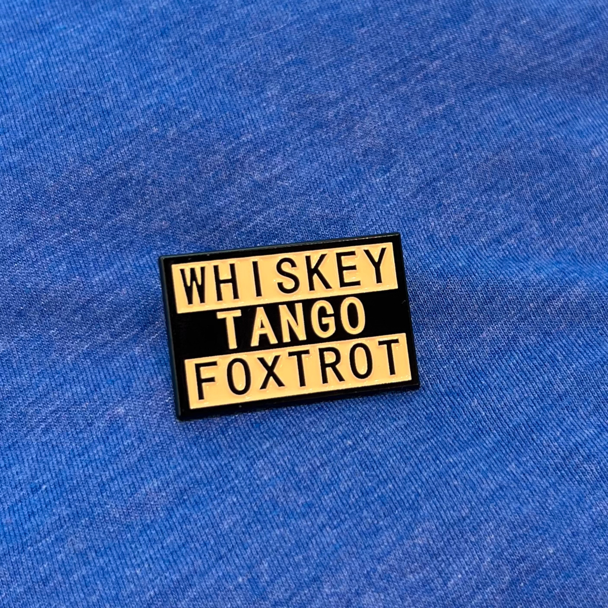 A rectangular enamel pin with a black background and yellow lettering that reads 'WHISKEY TANGO FOXTROT', placed on a blue textured fabric background.