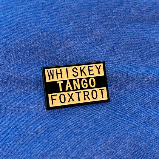 A rectangular enamel pin with a black background and yellow lettering that reads 'WHISKEY TANGO FOXTROT', placed on a blue textured fabric background.