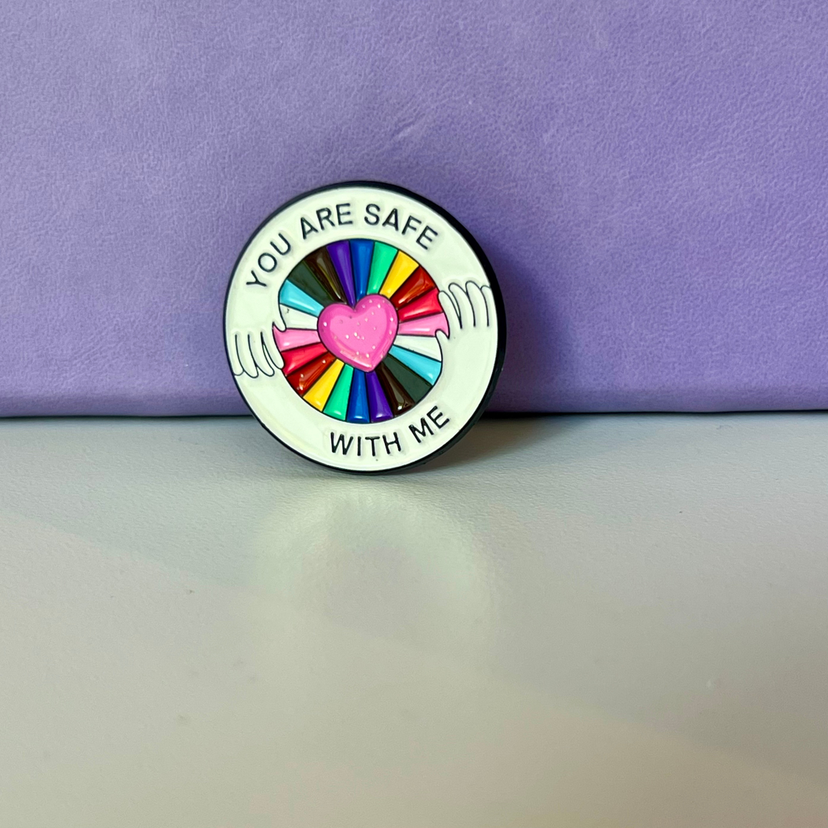 A round enamel pin with a white background and black border, featuring the text 'YOU ARE SAFE WITH ME' around a central design of a pink heart surrounded by rainbow-colored segments and white wing-like lines. The pin is photographed standing upright on a light gray surface against a textured purple background.