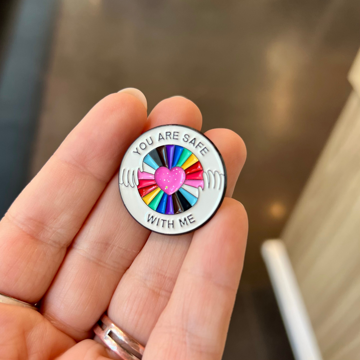 A close-up of a hand holding a round enamel pin that reads 'YOU ARE SAFE WITH ME' around a central pink heart and rainbow-colored hand motif.