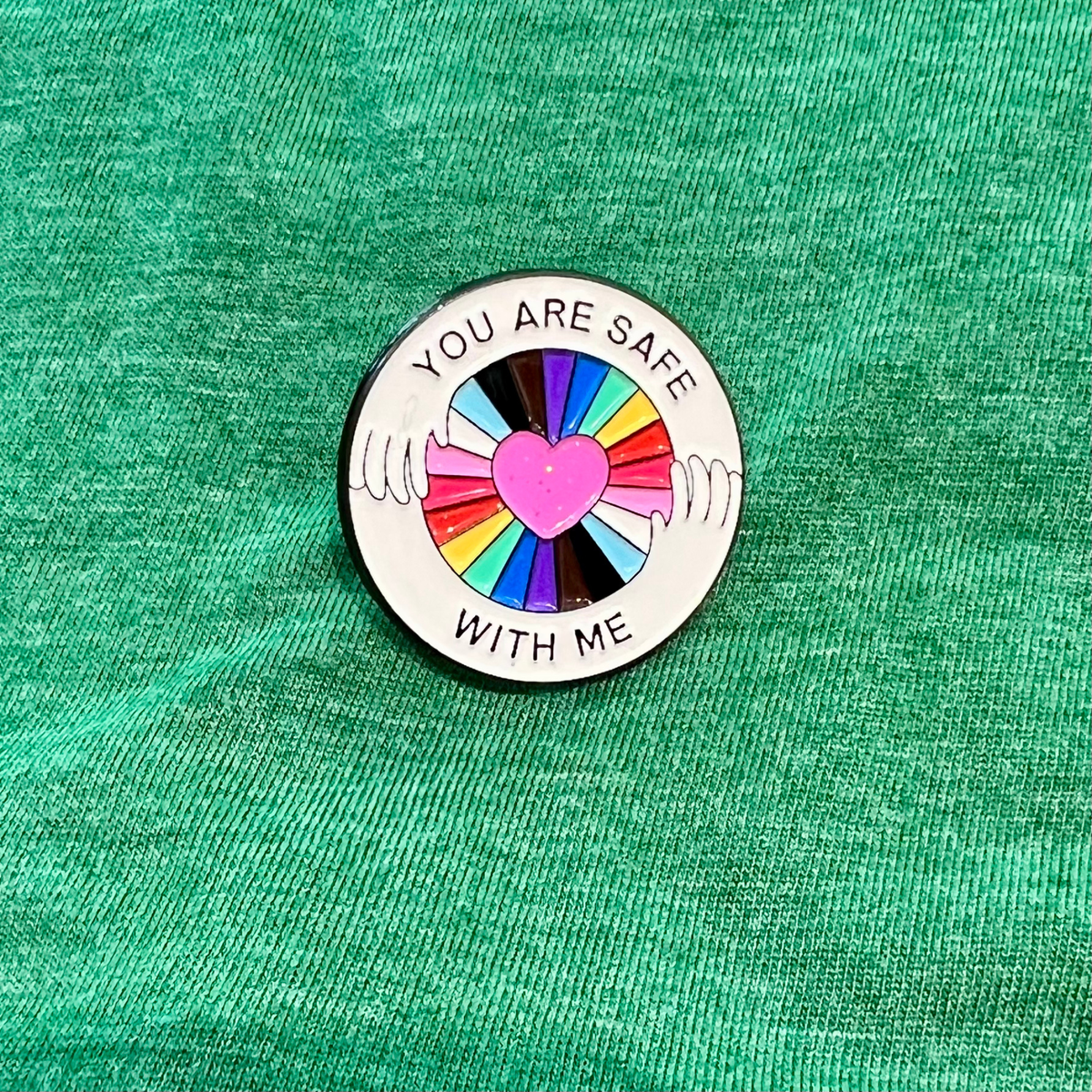 A circular enamel pin with the text 'YOU ARE SAFE WITH ME' surrounding a central pink heart and multicolored hand graphic, placed on a textured green fabric background.