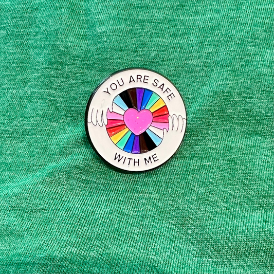 A circular enamel pin with the text 'YOU ARE SAFE WITH ME' surrounding a central pink heart and multicolored hand graphic, placed on a textured green fabric background.