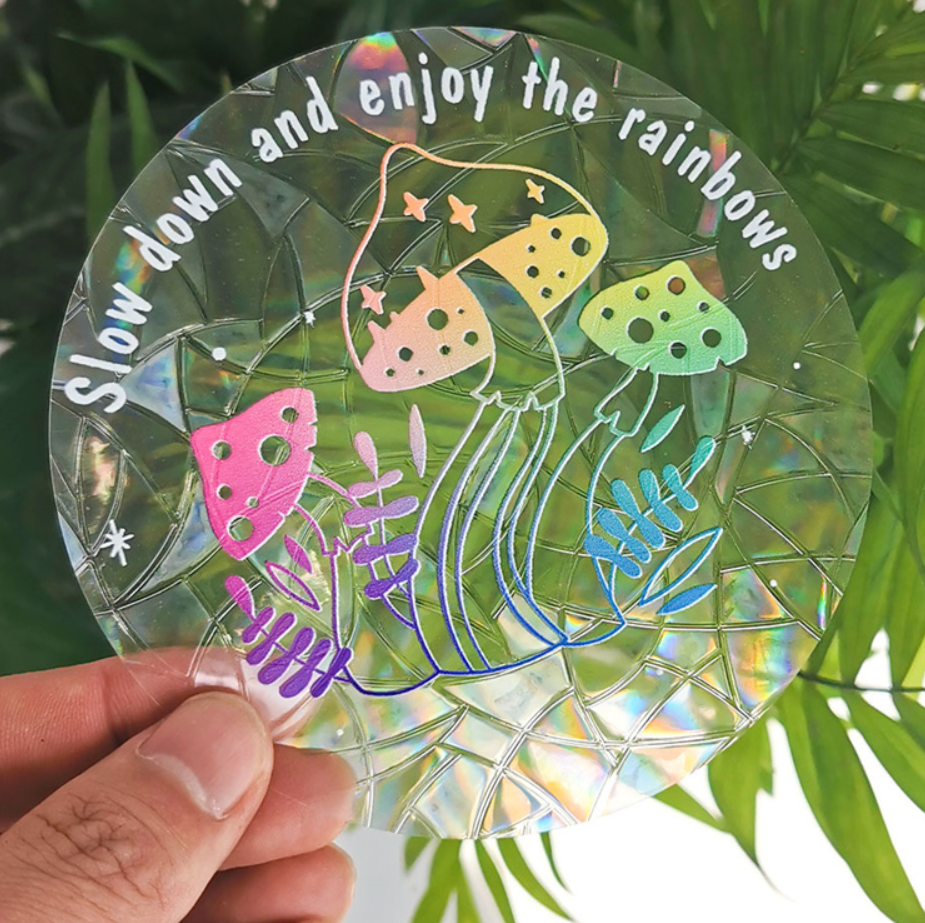 A hand holds the Mushroom Suncatcher by RainBee Designs LLC—a circular, iridescent window decal adorned with colorful mushrooms and ferns. The text reads, “Slow down and enjoy the rainbows,” with green leaves visible in the background.