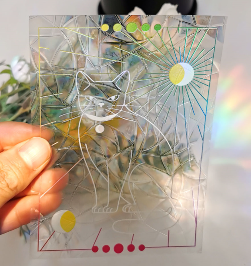 A hand holds the Cat Suncatcher by RainBee Designs LLC—a translucent card adorned with a white line drawing of a cat, geometric patterns, suns, and decorative colored circles—capturing the whimsical charm of a rainbow window decal, with plants and soft rainbow light in the background.